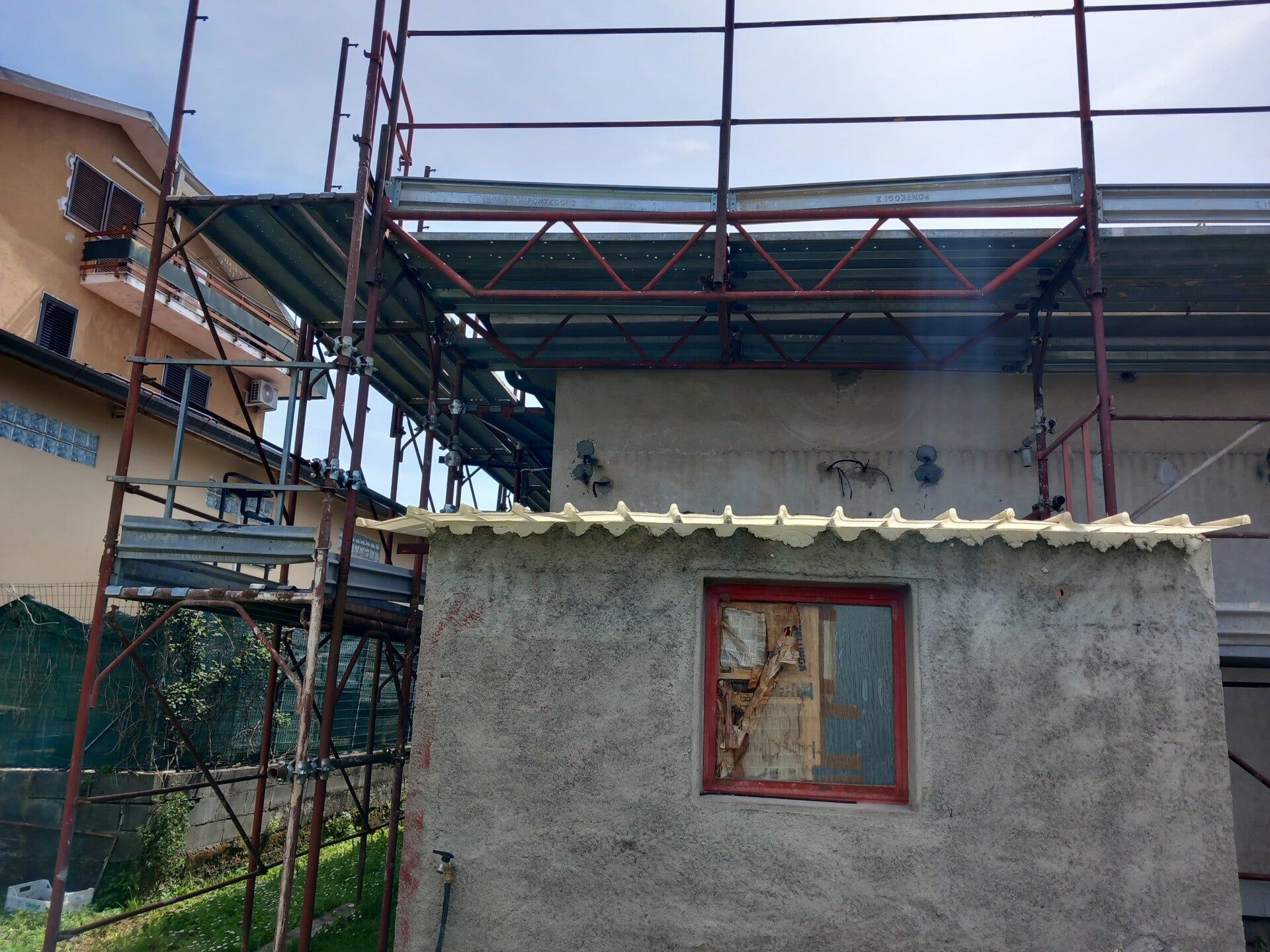 Casa parzialmente costruita con impalcature e una piccola dependance grigia con una finestra con cornice rossa.