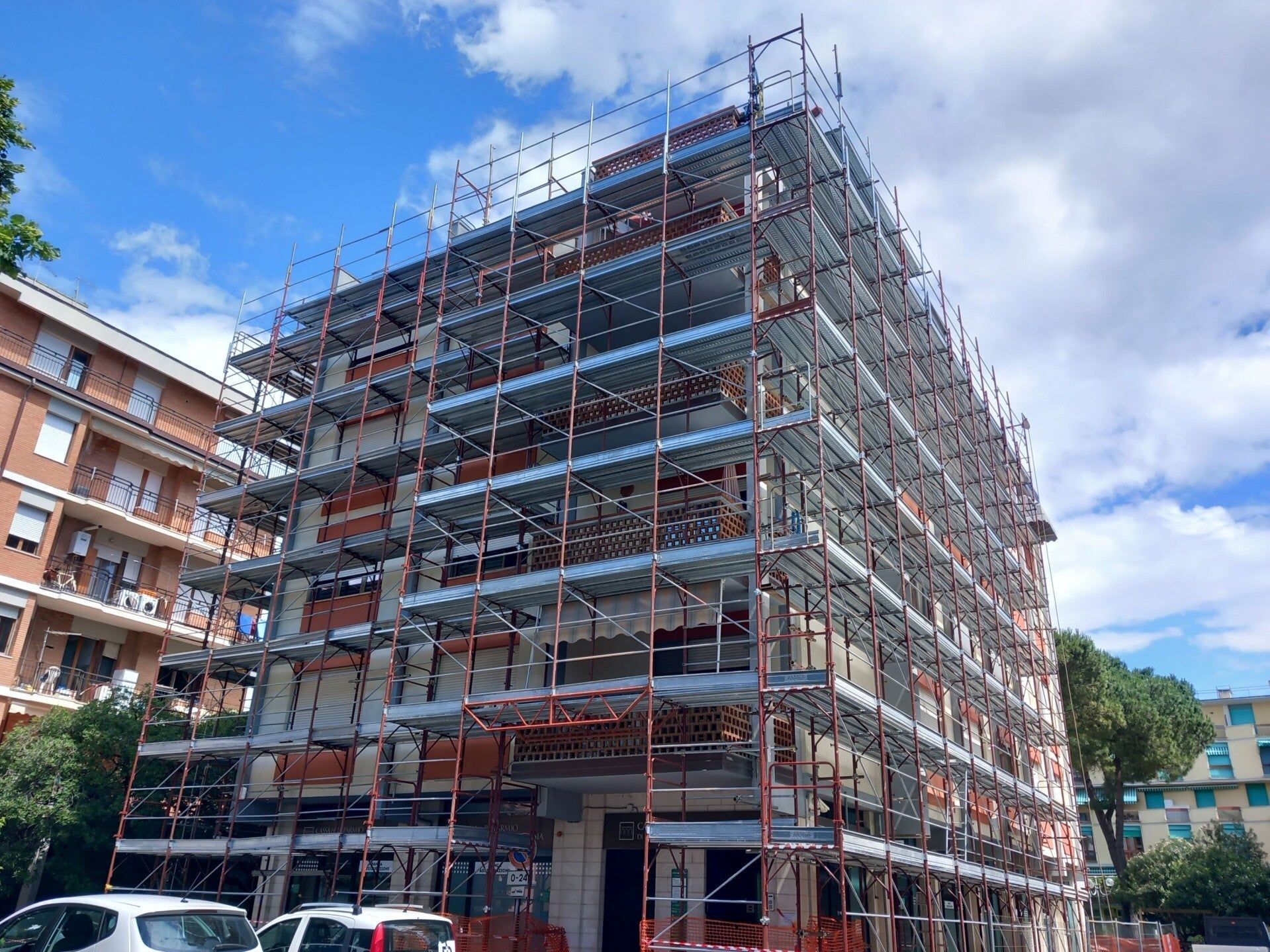 Edificio multipiano in costruzione, con impalcature, accanto a un condominio in una strada soleggiata.
