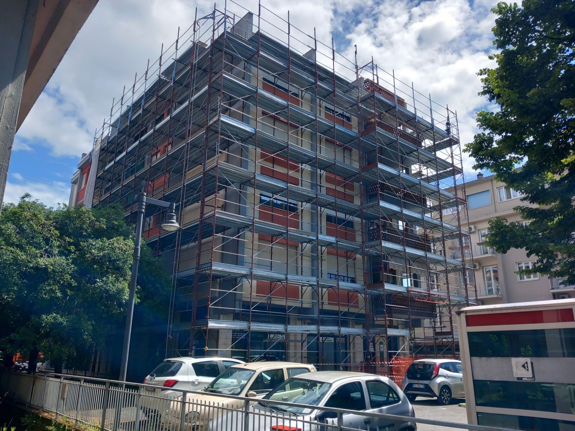 Impalcature che circondano un edificio a più piani in costruzione su una strada cittadina, con auto parcheggiate e alberi nelle vicinanze.