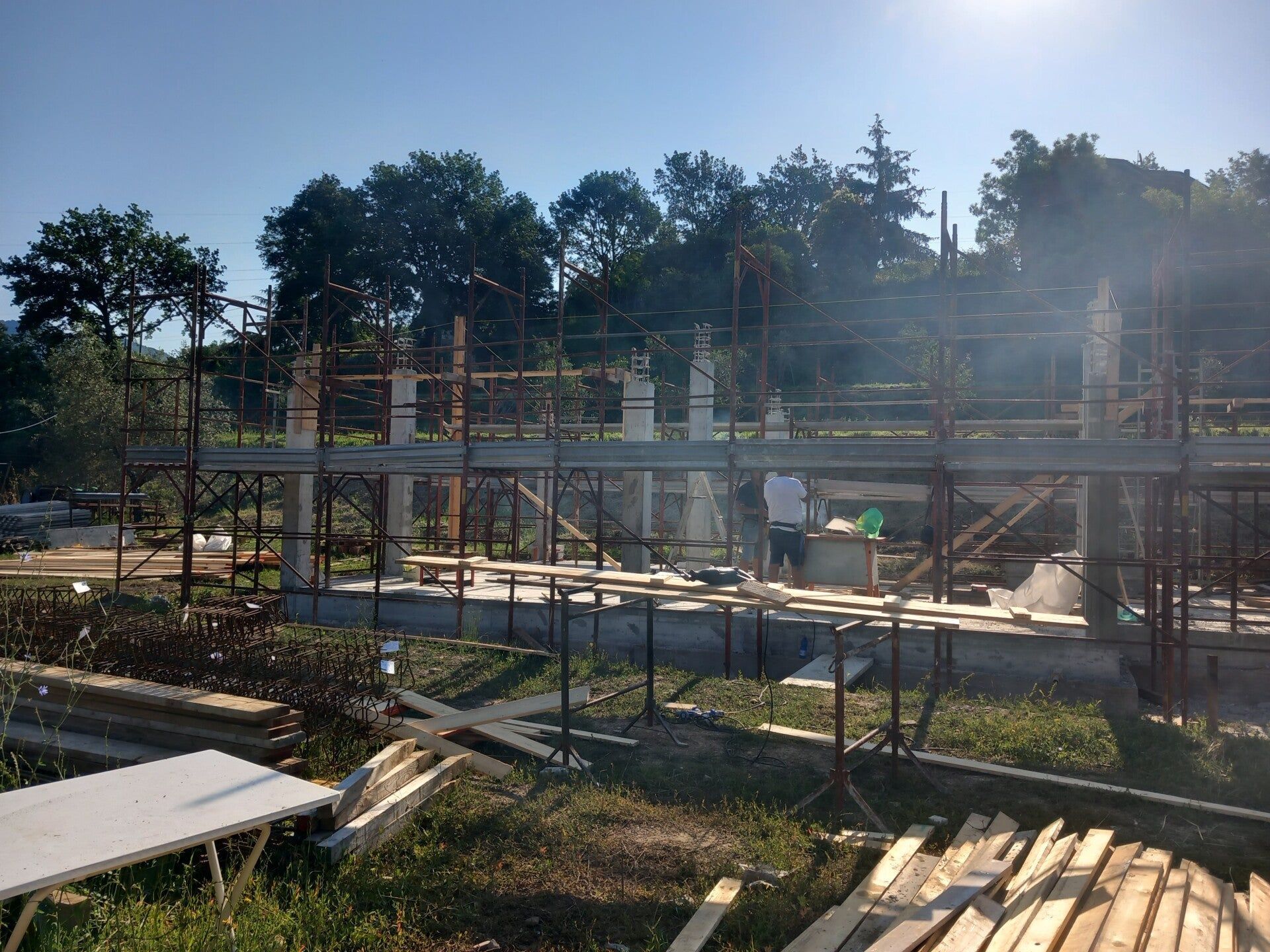 Cantiere edile con struttura in acciaio, impalcature e alberi sullo sfondo, sotto un cielo luminoso.