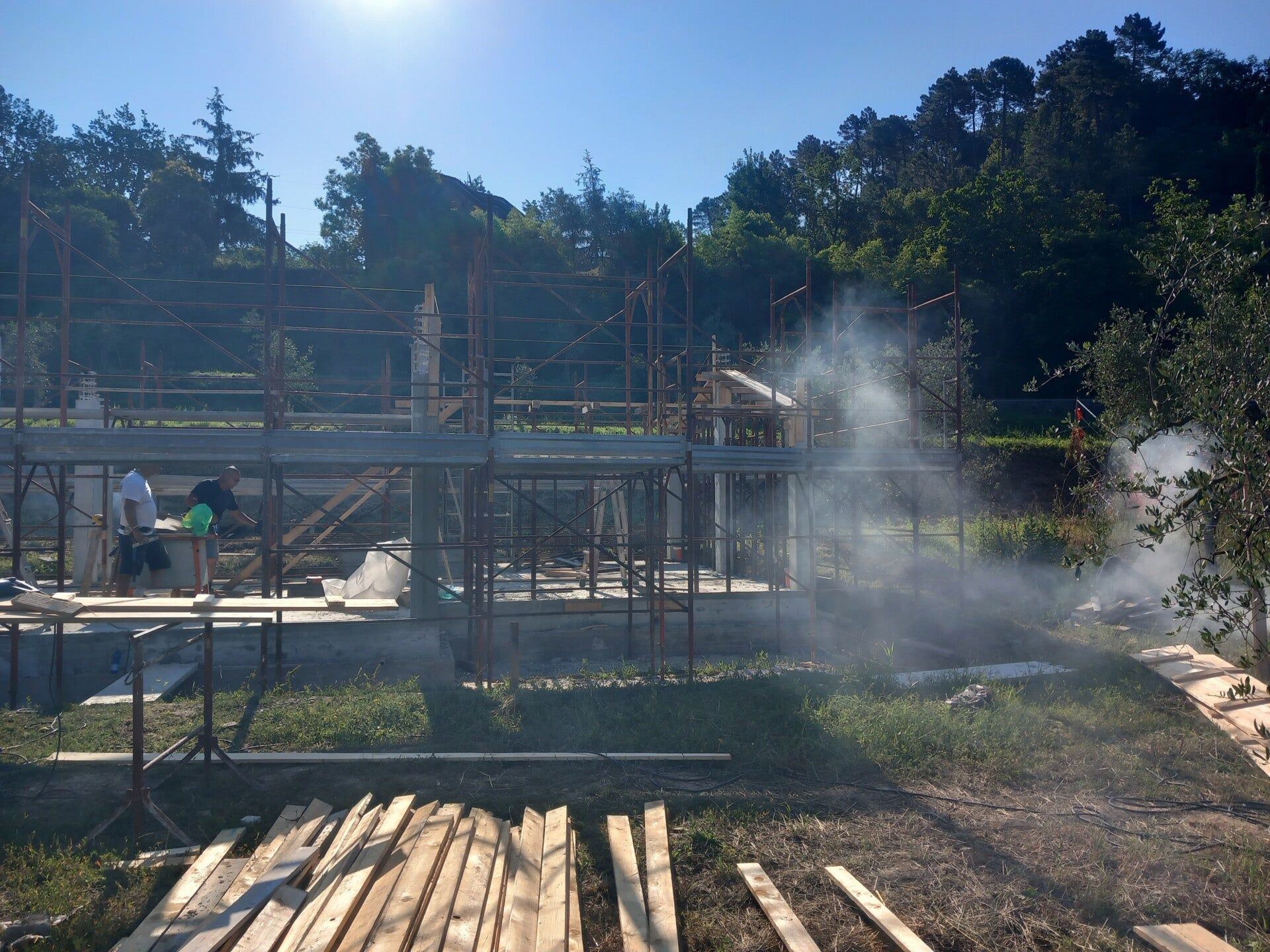 Cantiere edile con impalcature, operai e polvere che si alza sotto la luce intensa del sole.