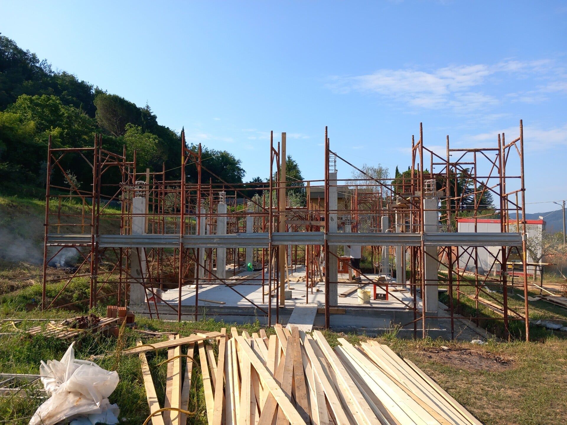 Cantiere edile con impalcature attorno a una struttura in cemento e legname accatastato in primo piano.