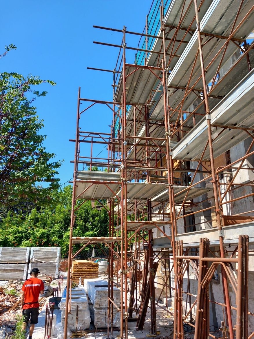 Cantiere edile con impalcature accanto a un edificio, un operaio in arancione vicino a materiali accatastati sotto un cielo azzurro.
