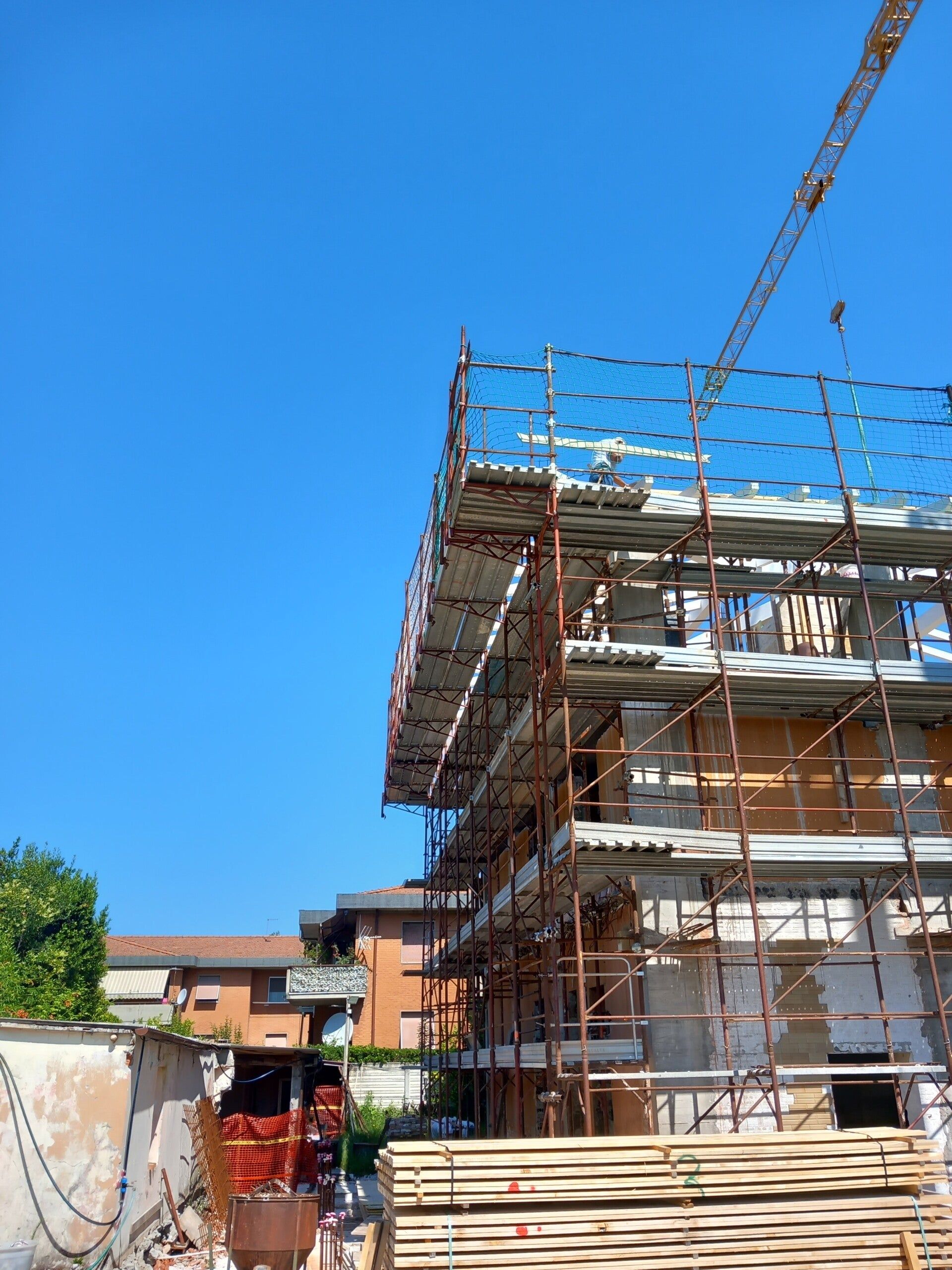 Impalcature attorno a un edificio in costruzione, accanto a cataste di legname in una giornata di sole.