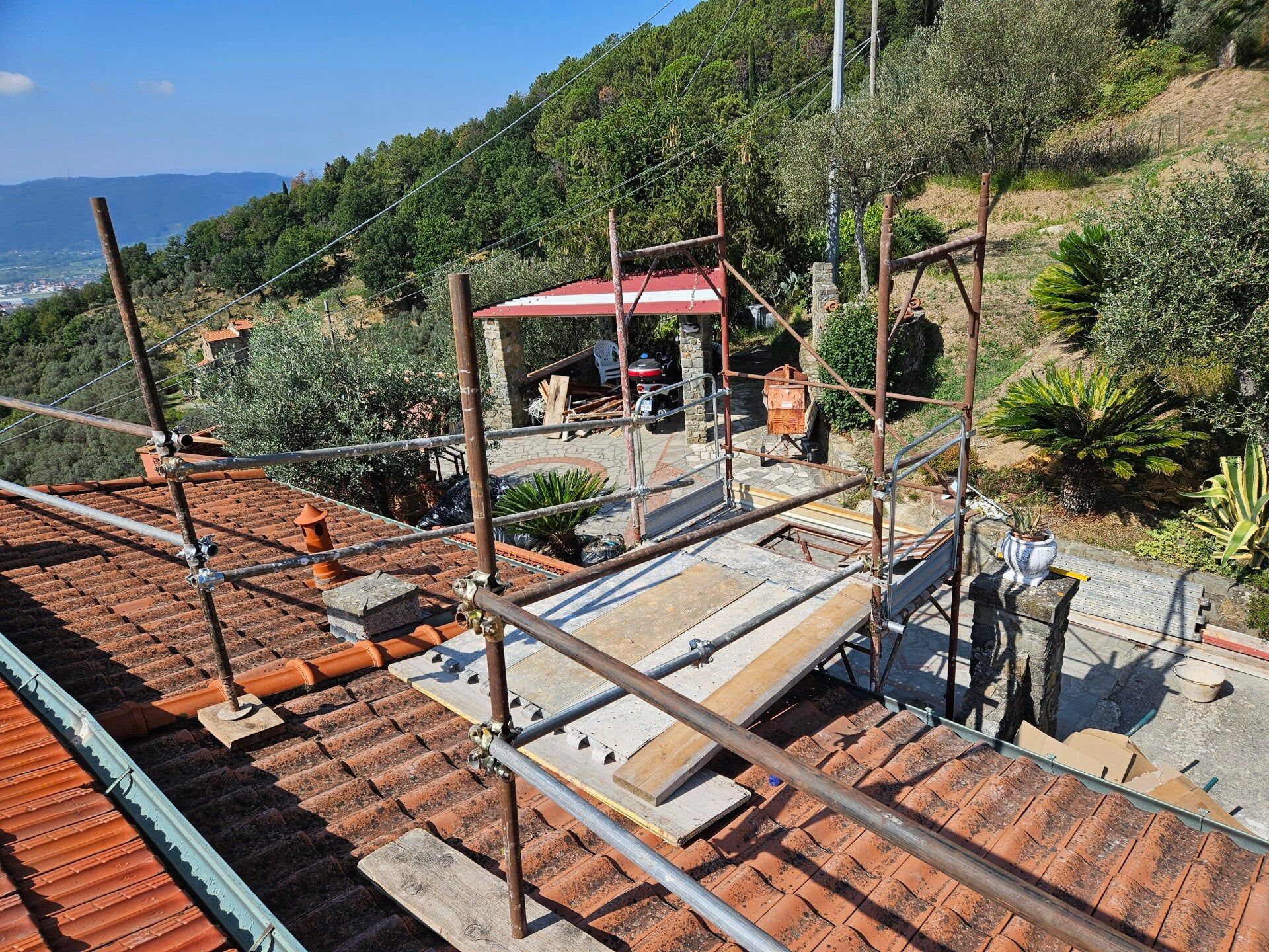 Lavori di costruzione sul tetto con tegole rosse, impalcature e operai accanto a una casa in collina sotto un cielo limpido.