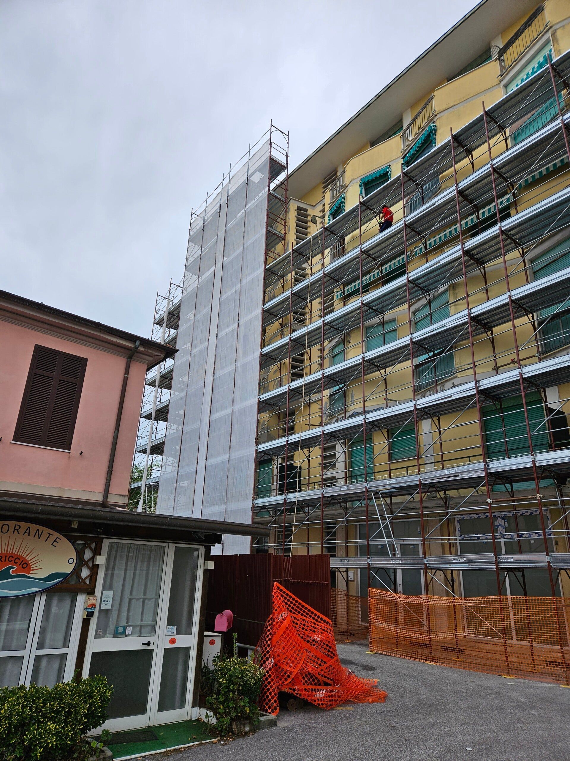 Un condominio in costruzione, con impalcature, si trova accanto a una piccola casa rosa, protetta da una rete arancione.