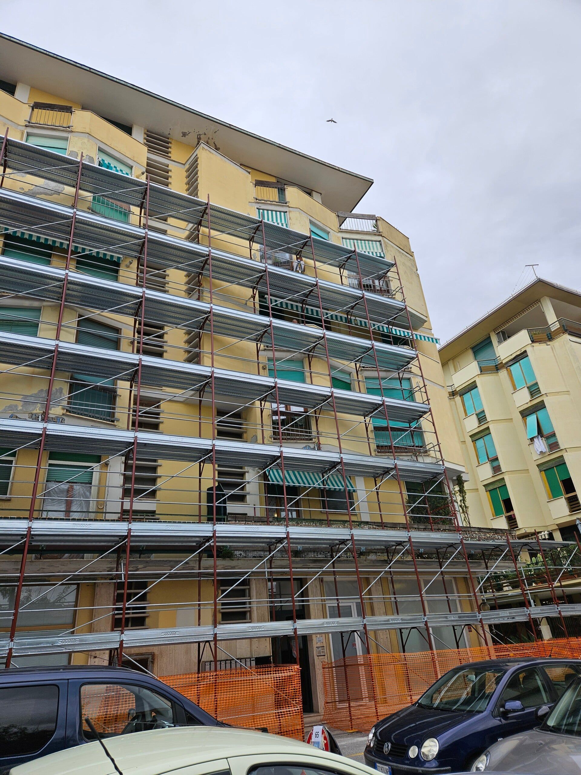 Impalcature attorno a un condominio giallo in costruzione, con auto parcheggiate davanti.