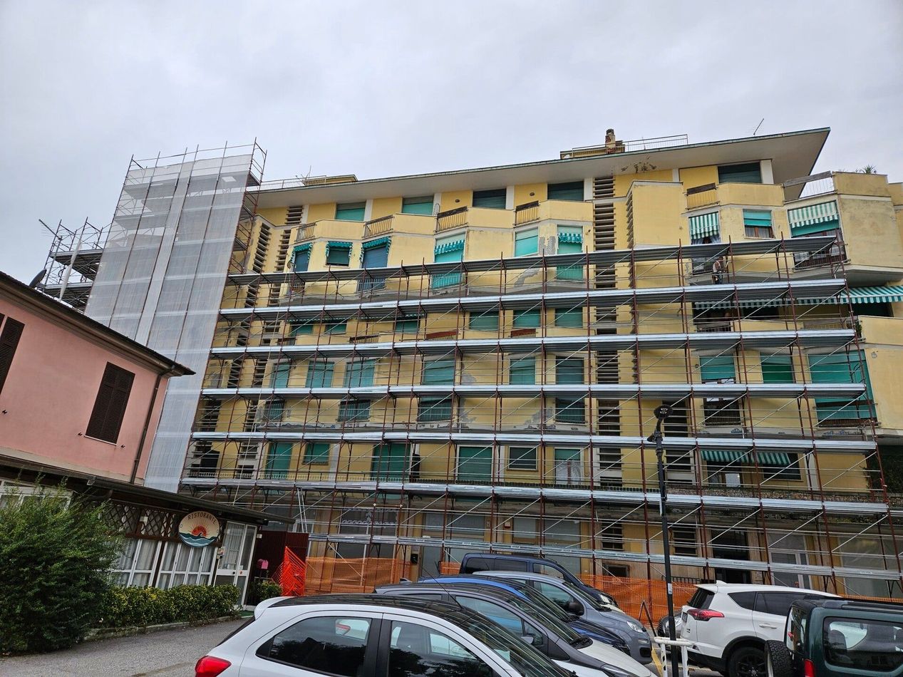 Palazzo di appartamenti giallo parzialmente impalcato, con auto parcheggiate davanti e cielo nuvoloso sopra.