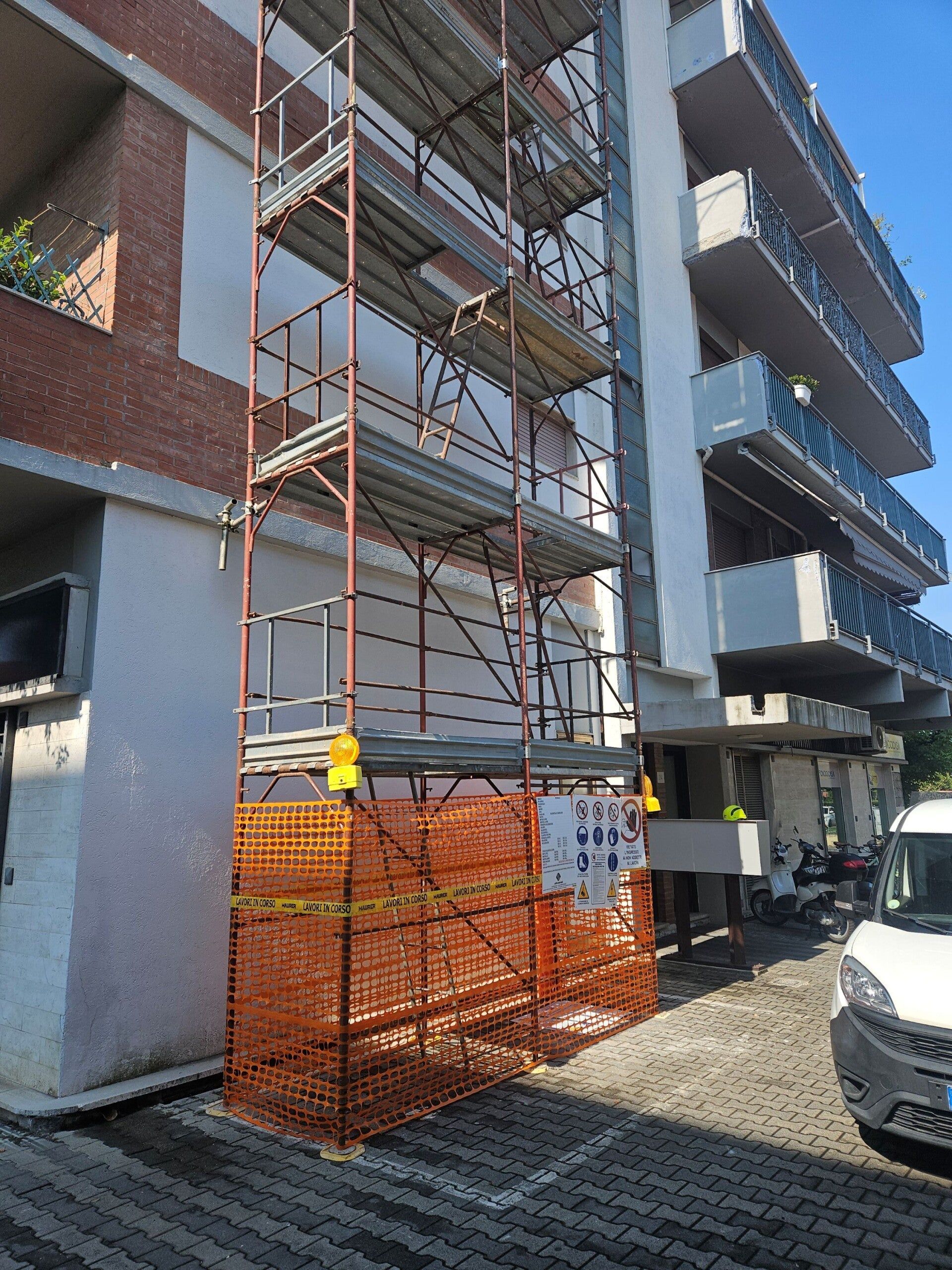 Ponteggi accanto a un condominio in mattoni, con recinzioni di sicurezza arancioni e un'auto parcheggiata nelle vicinanze.