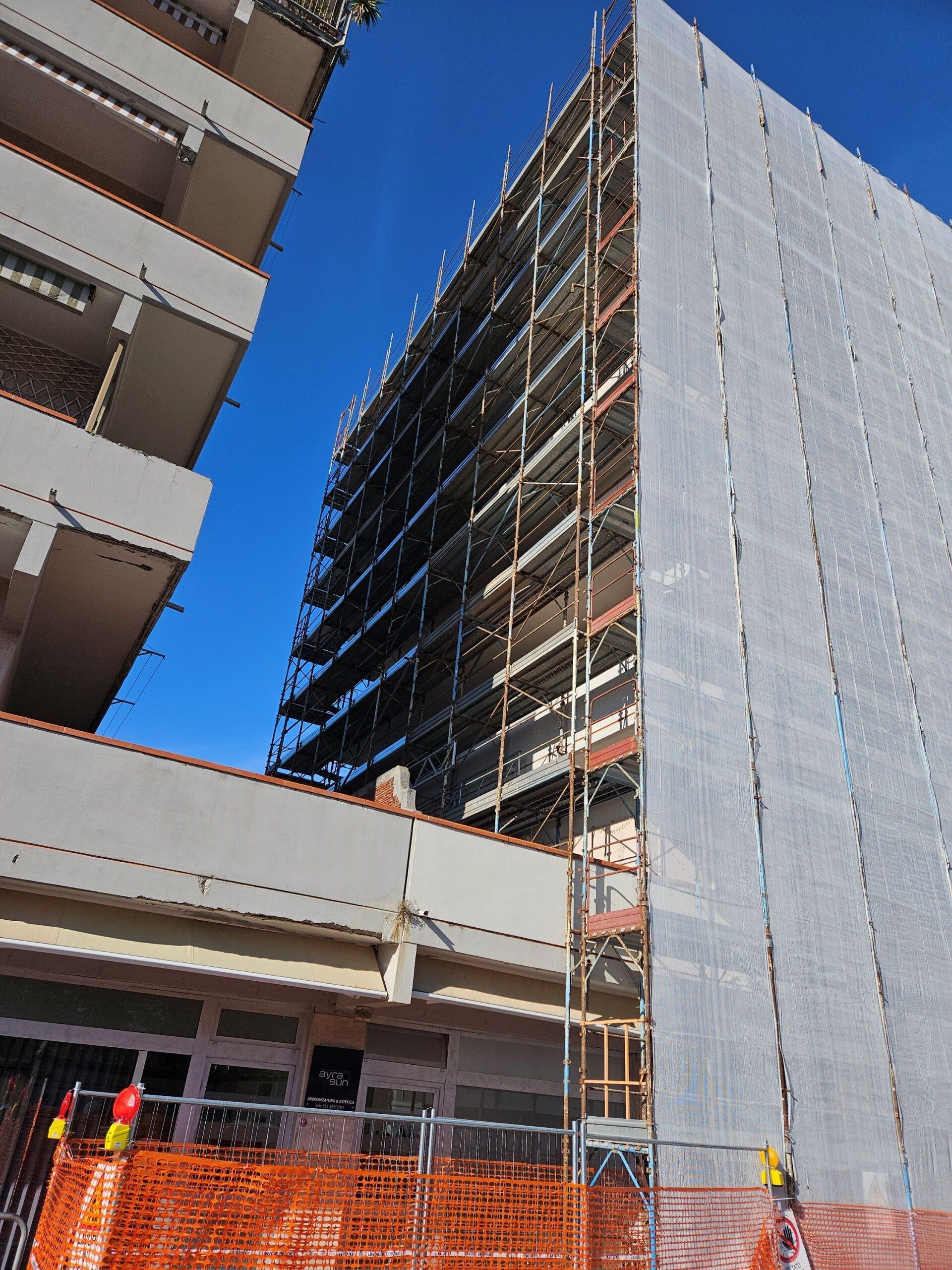 Edificio con impalcature in fase di ristrutturazione accanto a un'alta struttura avvolta in un telo grigio, sullo sfondo di un cielo azzurro.