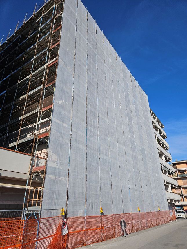 Cantiere edile con un edificio ricoperto di impalcature e reti grigie dietro una recinzione di sicurezza arancione sotto un cielo azzurro.
