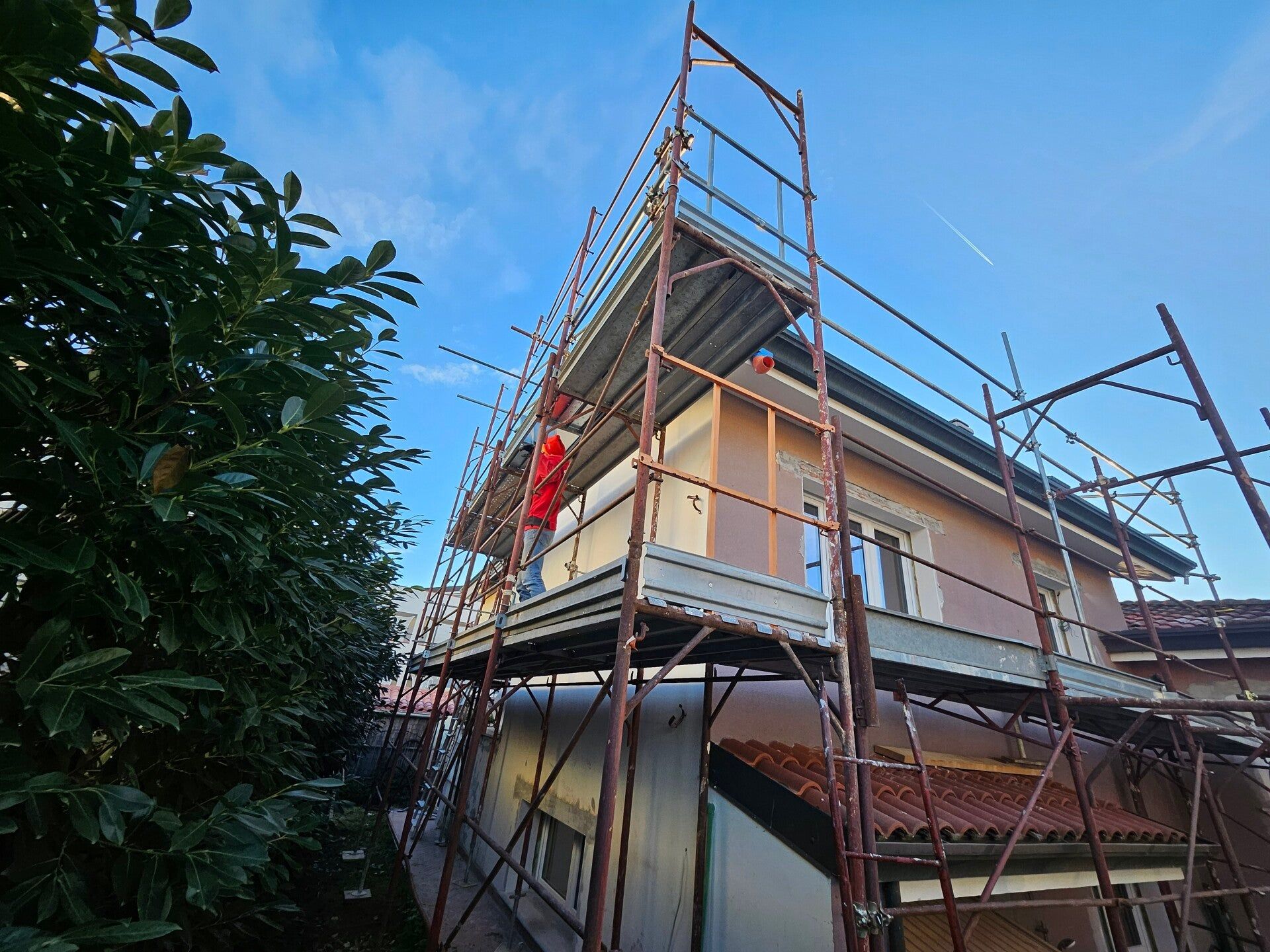 Casa in costruzione con impalcature e un operaio in divisa rossa al piano superiore.