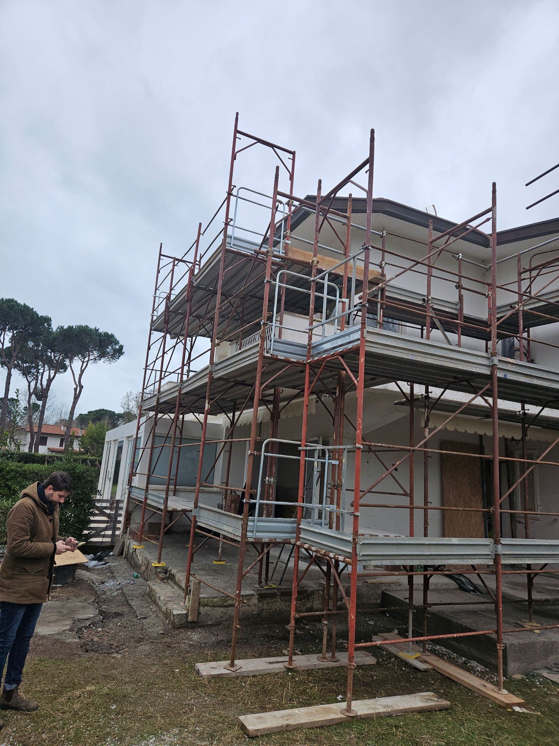 Casa in costruzione con impalcature metalliche e un operaio in piedi nelle vicinanze sotto un cielo nuvoloso.
