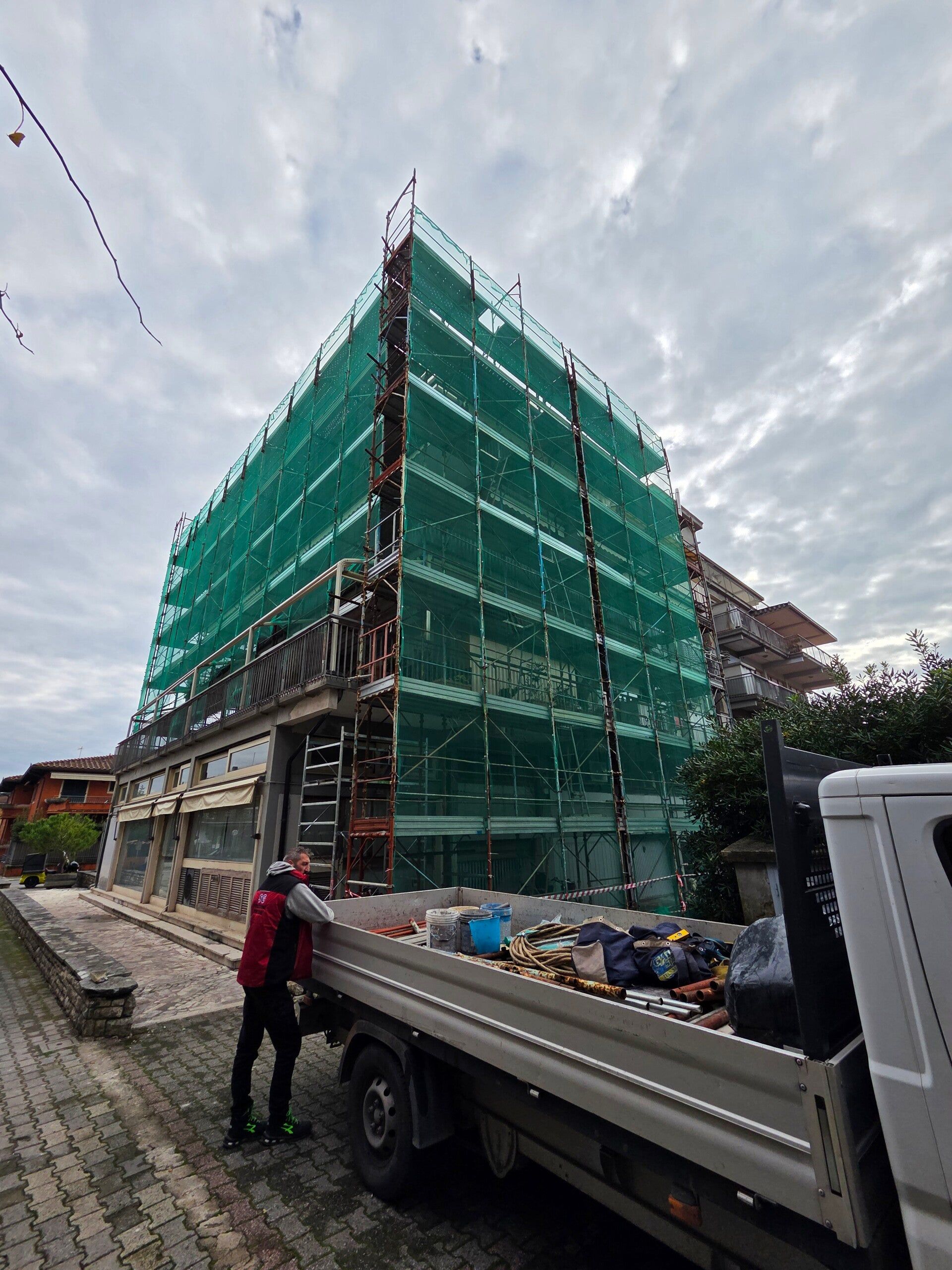Cantiere edile con una struttura impalcata coperta da una rete verde e un camion a pianale davanti, sotto un cielo nuvoloso.