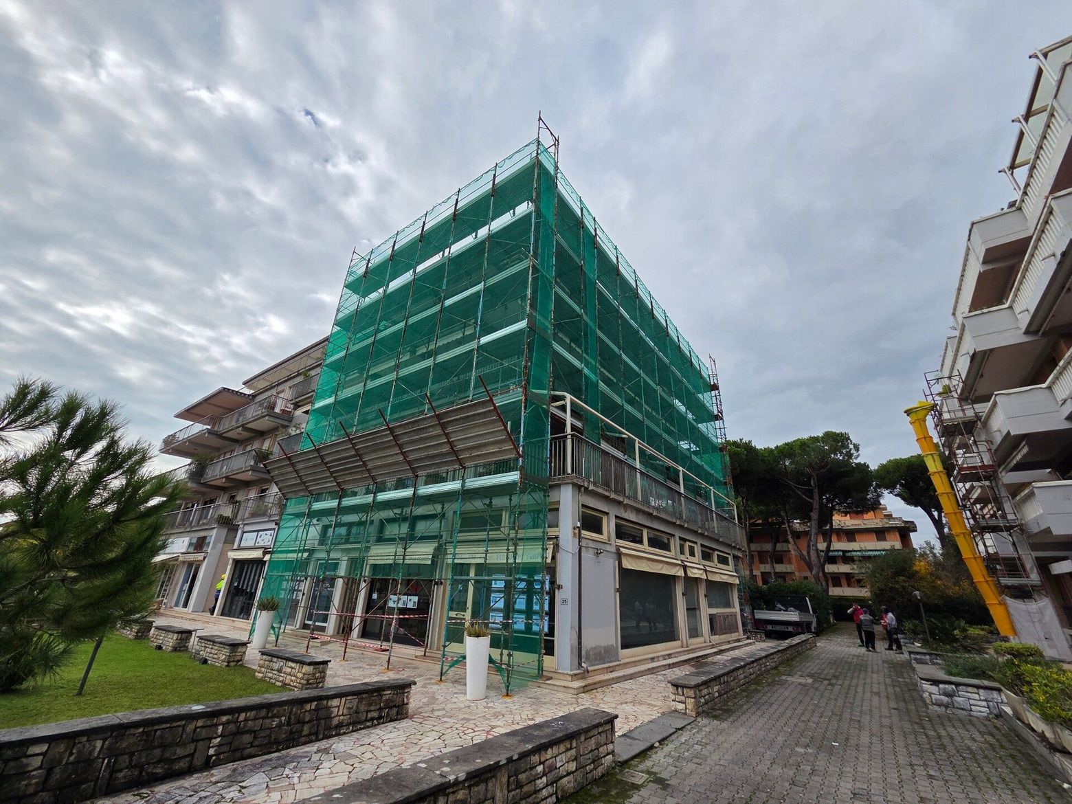 Edificio in costruzione ricoperto da una rete verde, situato su una strada nuvolosa, con case vicine e un marciapiede bagnato.