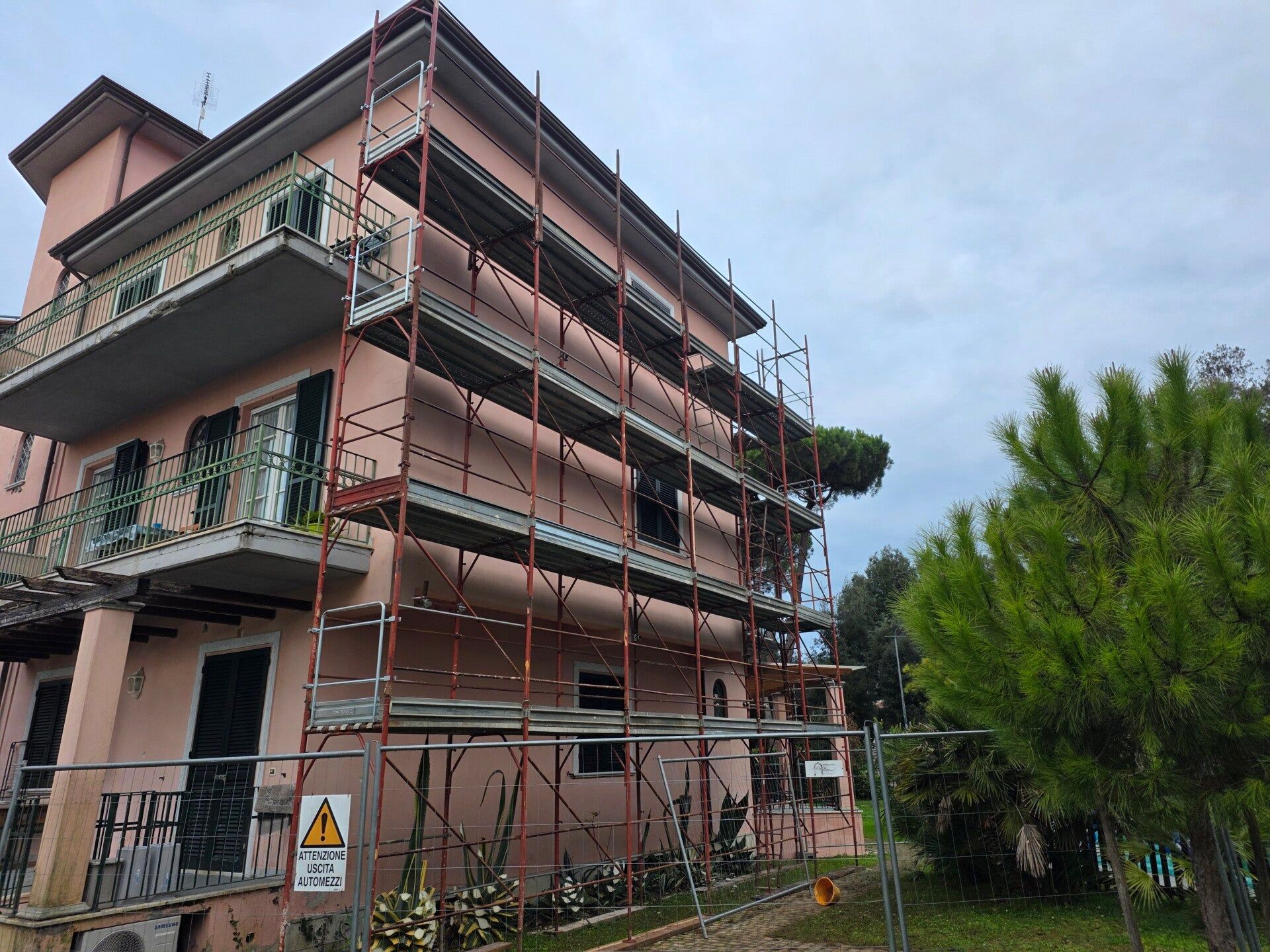 Edificio rosa in costruzione con impalcature accanto a un albero verde e un cielo nuvoloso.