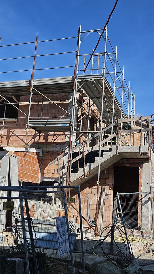 Casa in mattoni parzialmente costruita con impalcature e scala in cemento sotto un cielo azzurro
