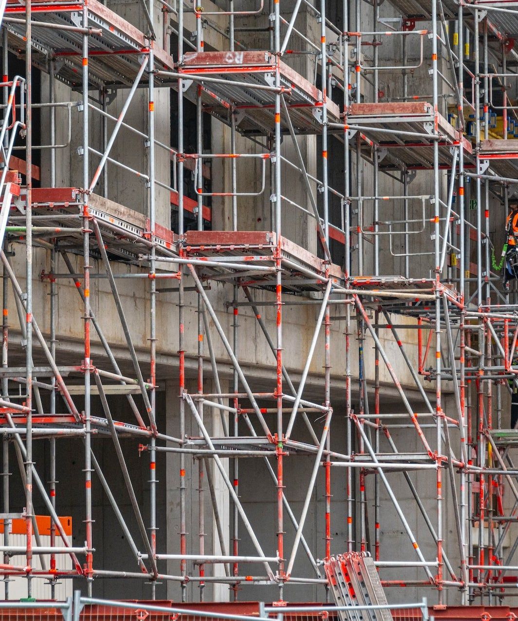 Impalcatura che ricopre la facciata di un edificio durante i lavori di costruzione, con piattaforme metalliche rosse e supporti grigi.