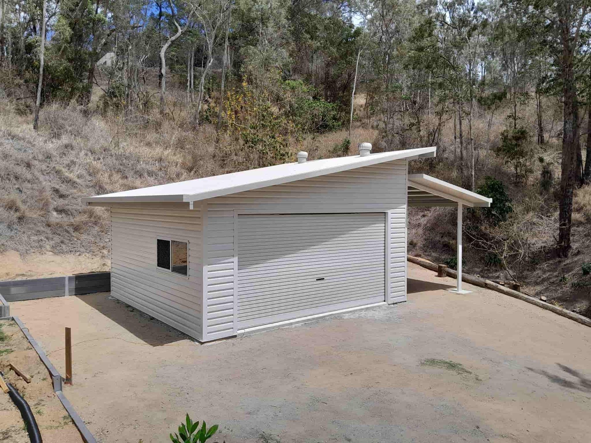 A white, single-story garage with a slanted roof and attached side carport, situated on a dirt lot near a wooded hill.