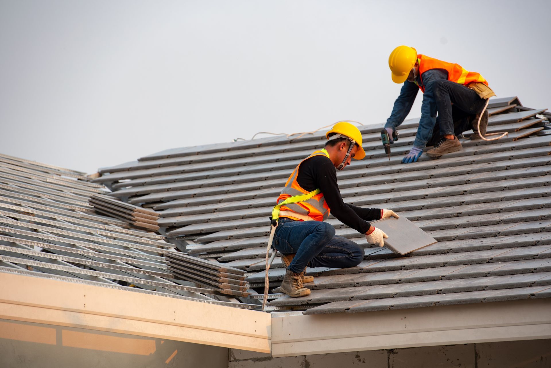 Two roofers in yellow hard hats installing gray roof tiles on a house under a cloudy sky.