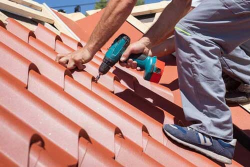 A man is working on a roof with a drill. Roofer using a drill to attach red roof tiles on a slanted roof under a bright sky.