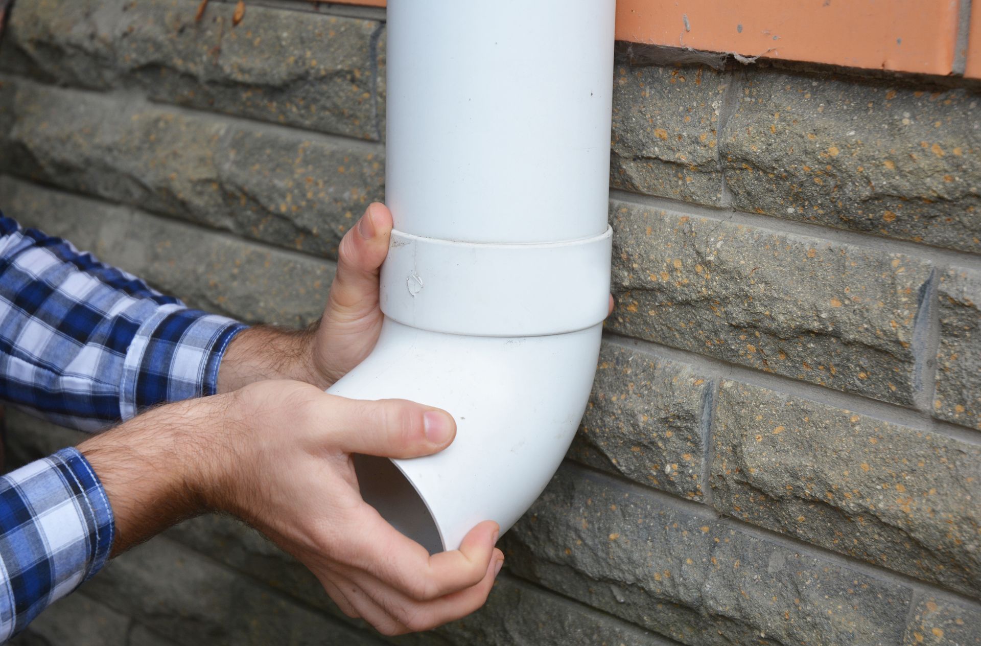 Close-up of a gutter contractor’s hands as he installs a PVC rain gutter drain pipe. Person fitting a white downspout to a brick wall.