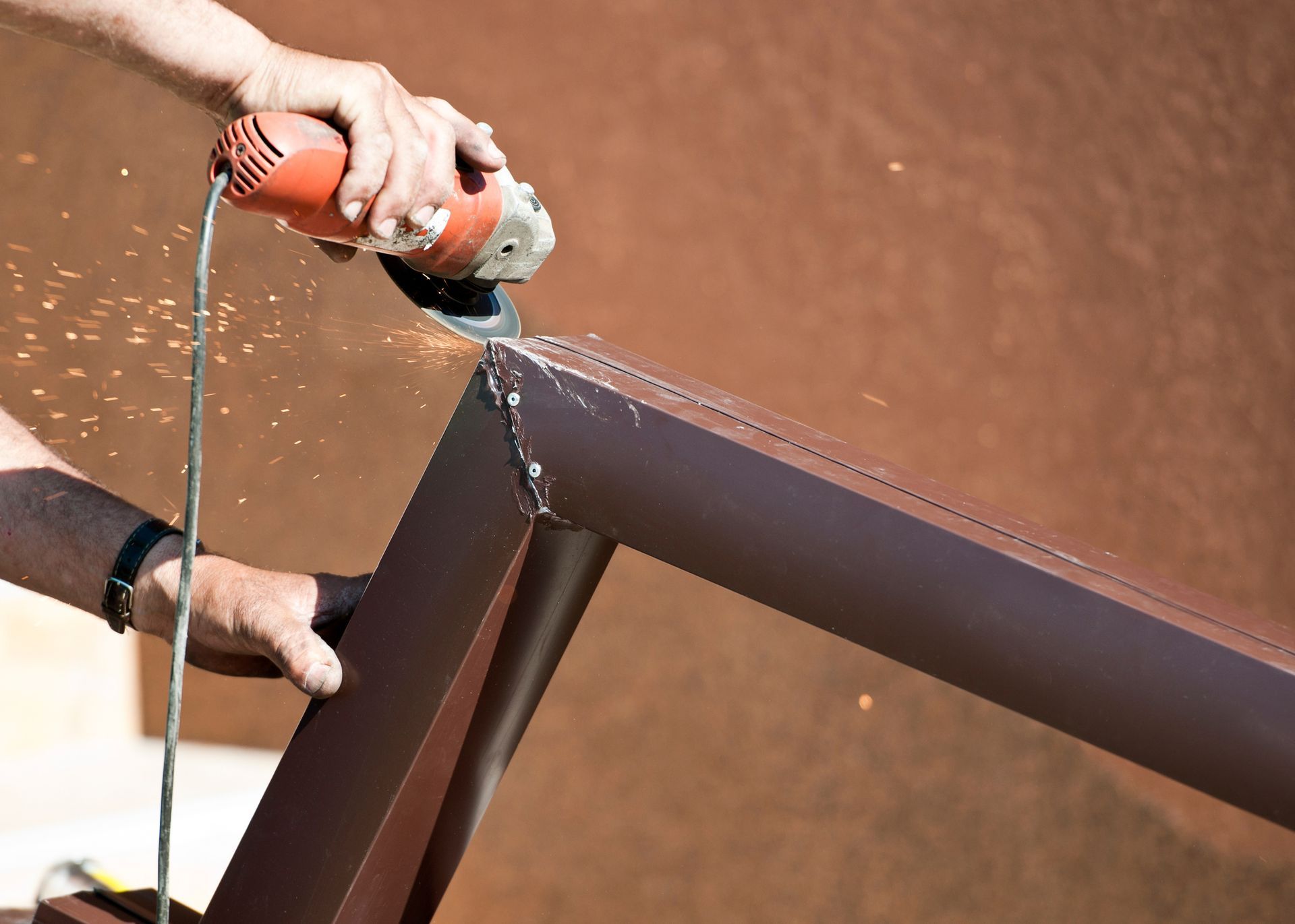 Person uses an angle grinder to cut a brown metal frame outdoors, creating sparks.
