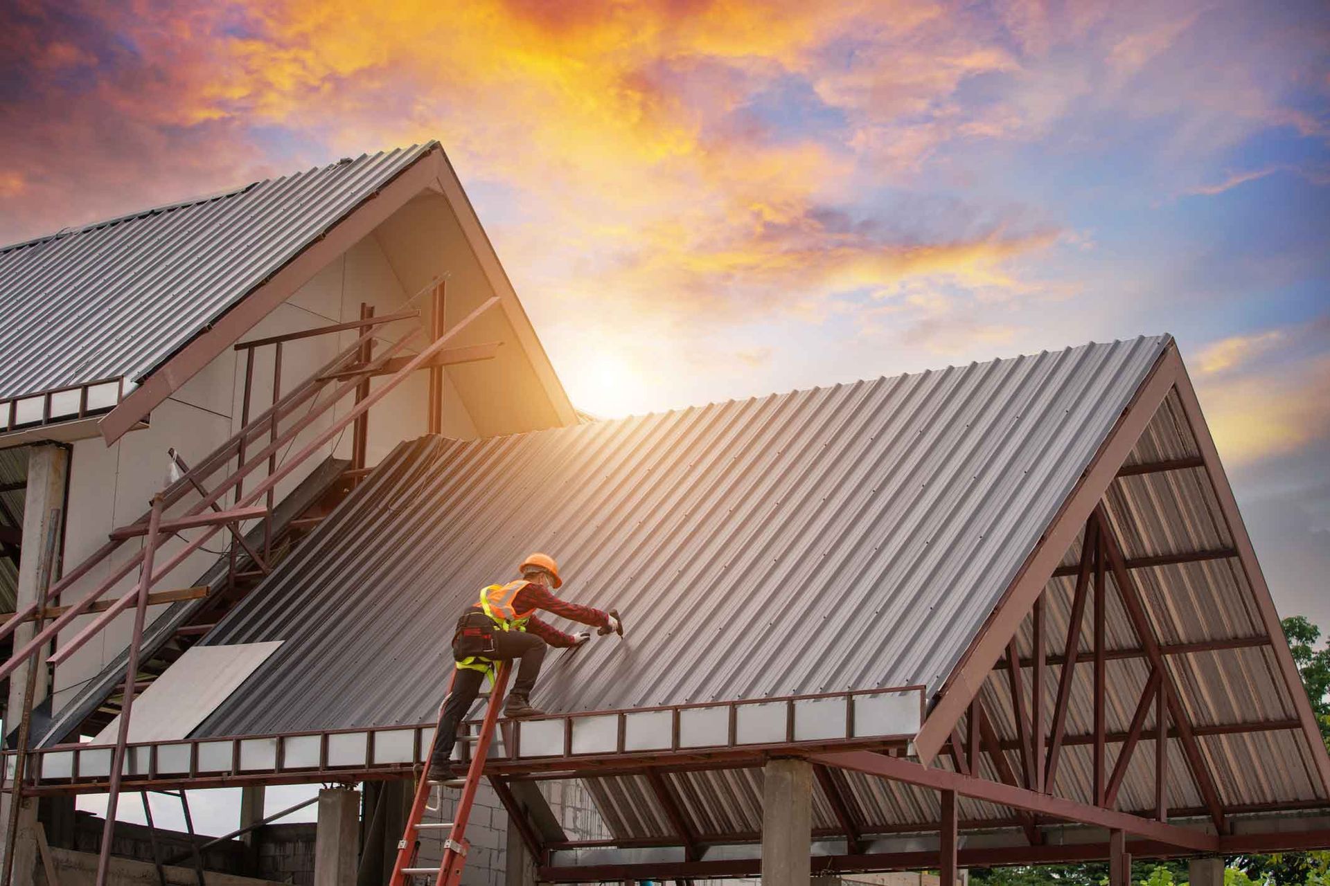 Roofer on a ladder installing metal roofing on a house under construction; sunset sky.