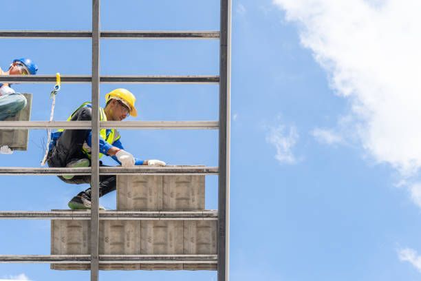 Construction worker in yellow hard hat and vest installing panel on building under blue sky.