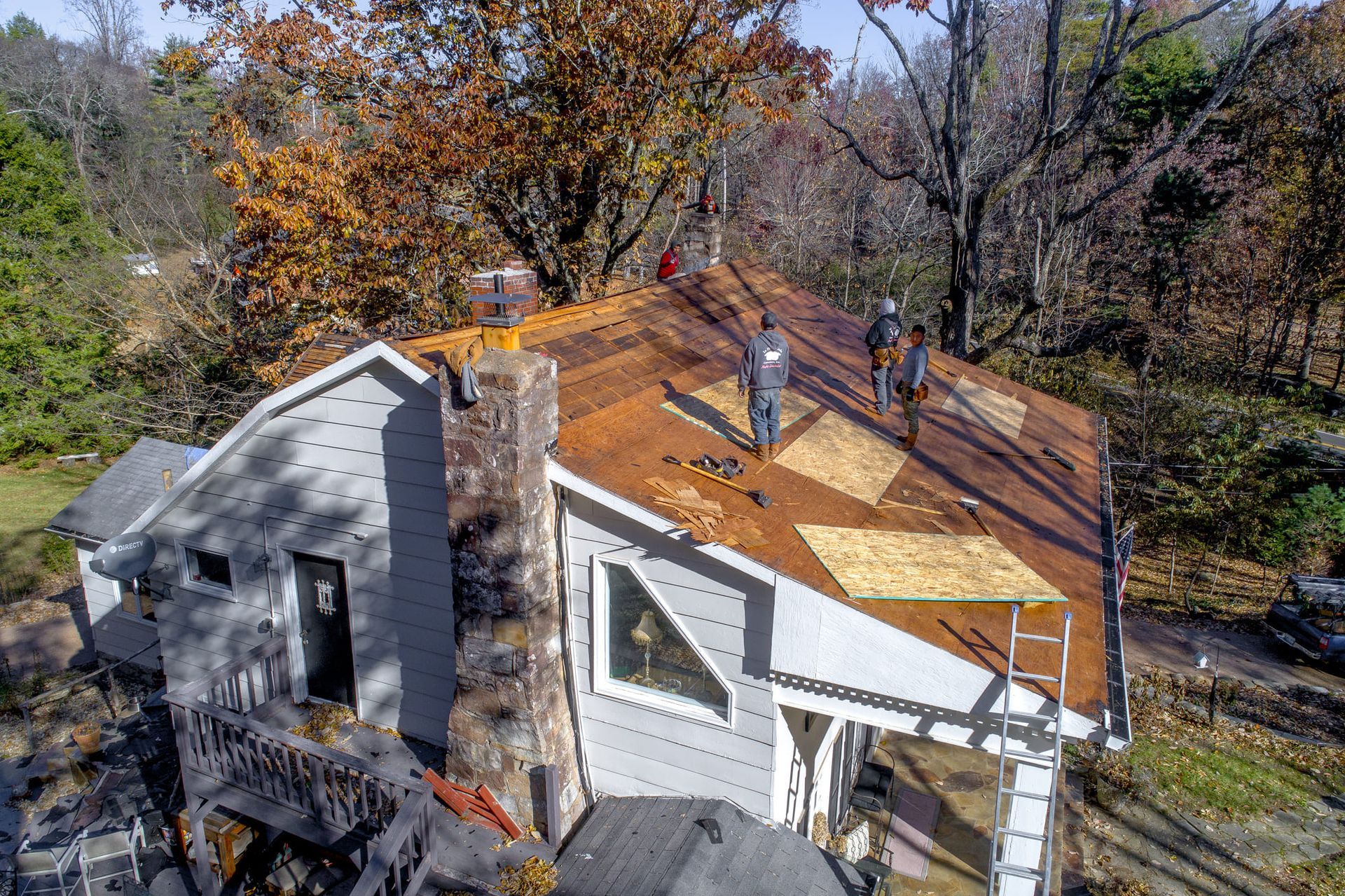 Three roofers work on a house roof. The roof is partially covered with wood. Autumn foliage surrounds.