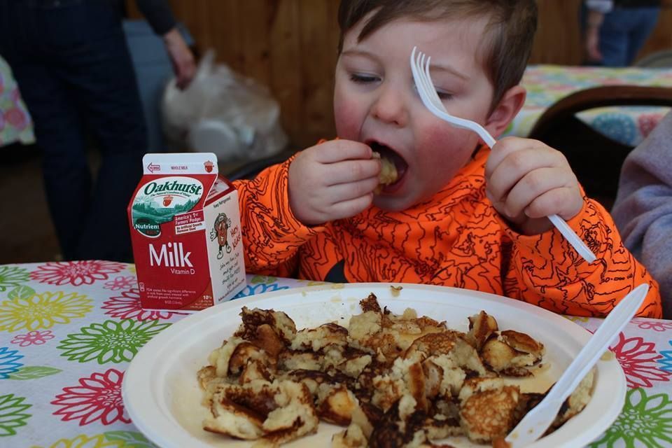 A child wearing an orange patterned hoodie eats small pieces of food from a white plate with a plastic fork.