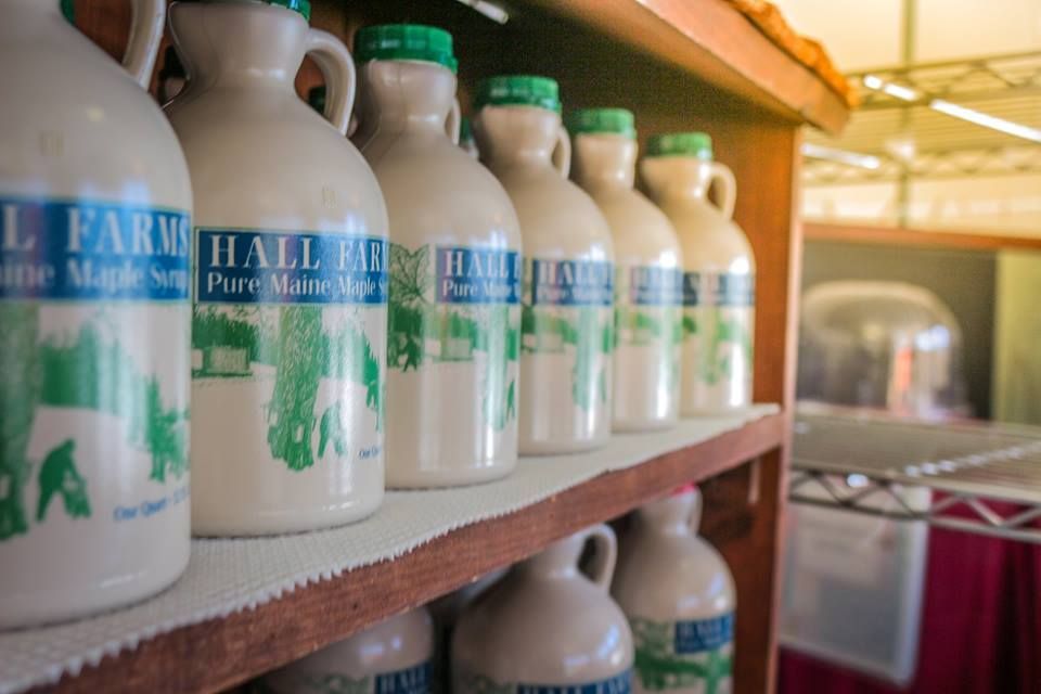 Ceramic jugs of Hall Farms maple syrup are neatly arranged on wooden shelves in a shop.