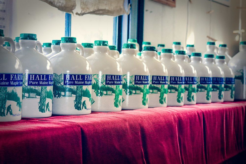 A row of white jugs of Hall Farm pure maple syrup lined up on a table covered by a red cloth.