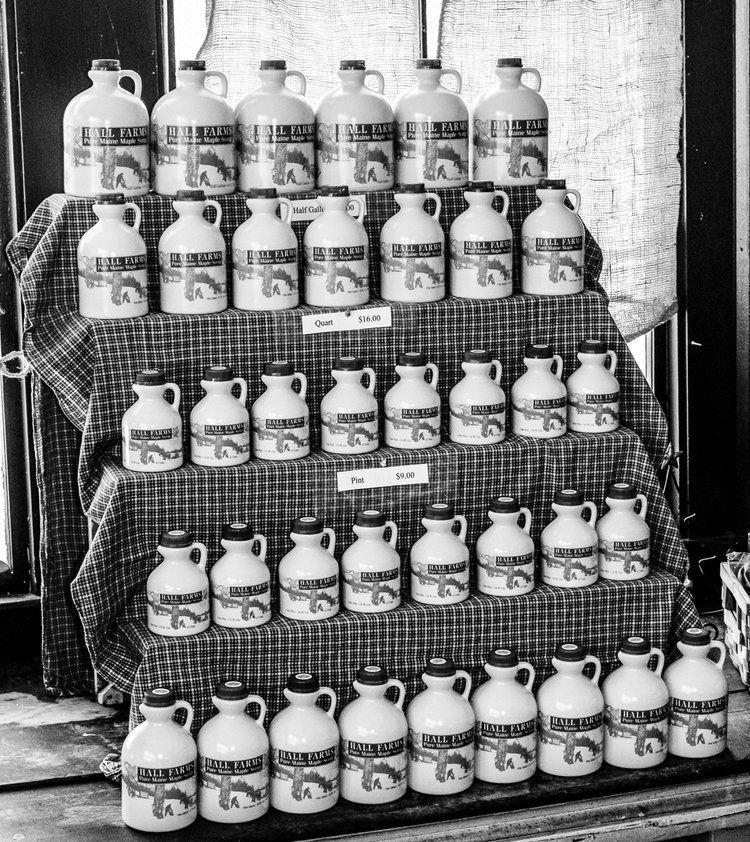 Five rows of identical jugs filled with maple syrup displayed on a tiered, fabric-covered shelf.