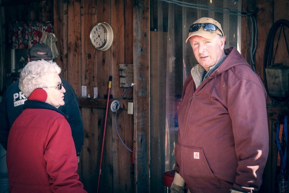 An older couple stands in a rustic wooden building, the man wearing a maroon jacket and cap, the woman in a red coat.