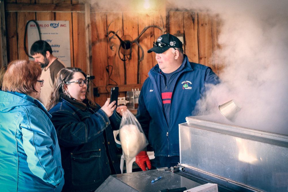 A person records with a phone as another person holds a plastic bag near a steaming vat inside a rustic wooden building.