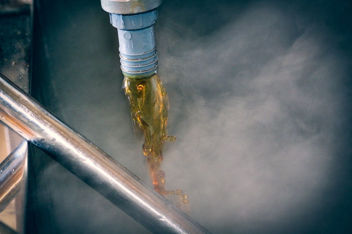 Golden liquid flows from a grey pipe into a steaming, dark industrial vat.