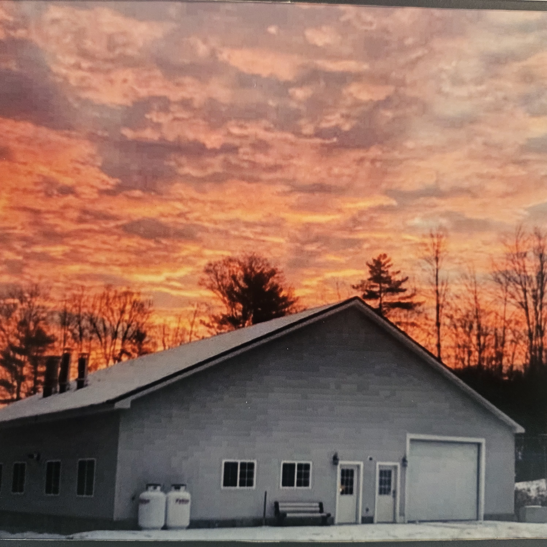 A gray workshop building stands against a vibrant orange and pink sunset sky with silhouetted trees in the background.