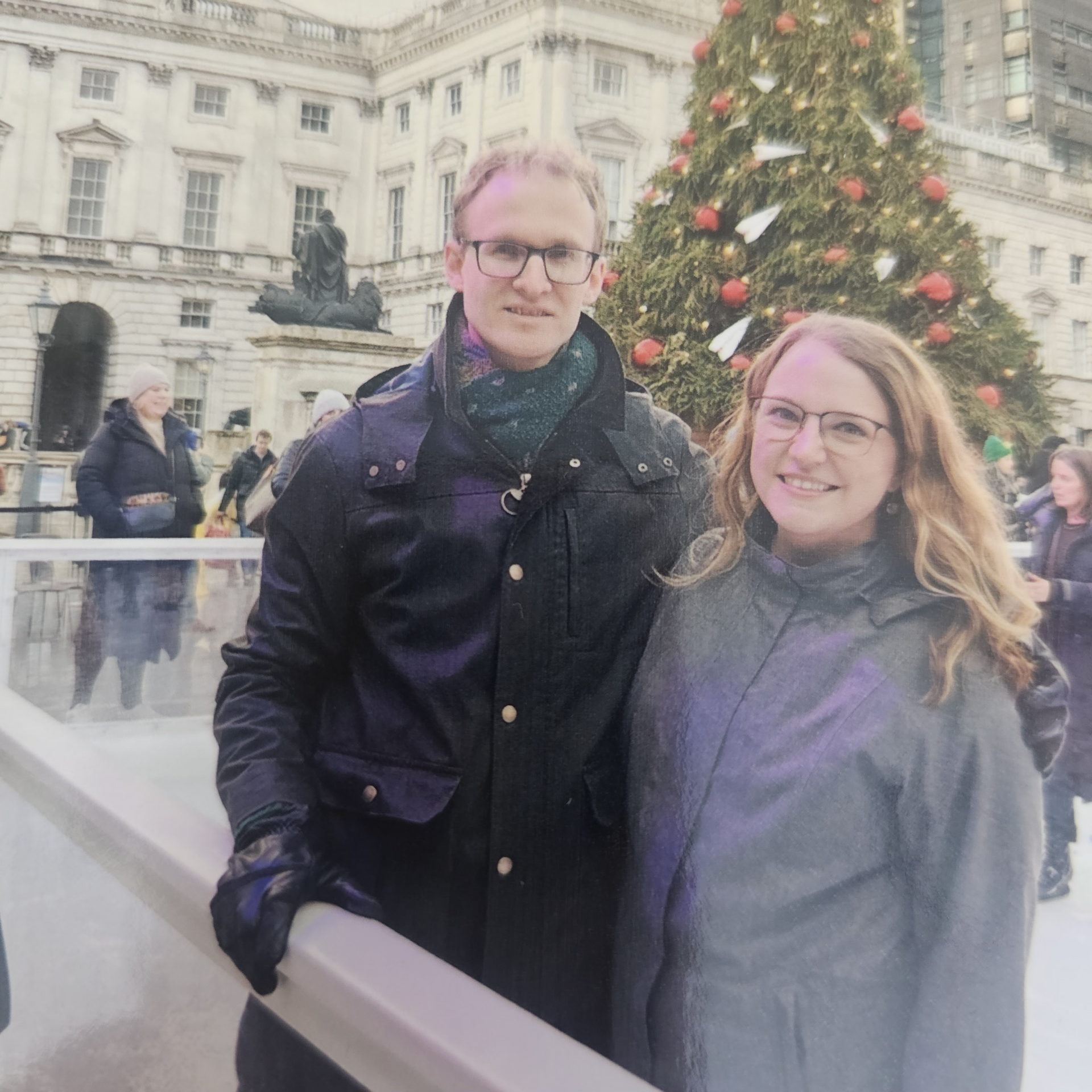 A smiling couple poses for a photo in front of a Christmas tree in a courtyard with neoclassical architecture.
