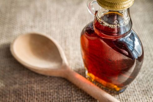 A glass bottle of dark amber maple syrup next to a wooden spoon, set against a textured burlap background.