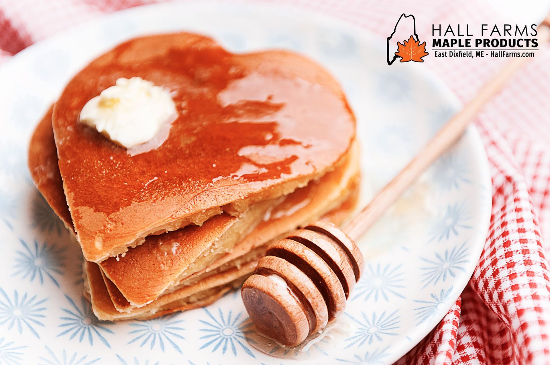 Heart-shaped pancakes topped with syrup and butter on a patterned plate, with a wooden honey dipper and red plaid cloth.