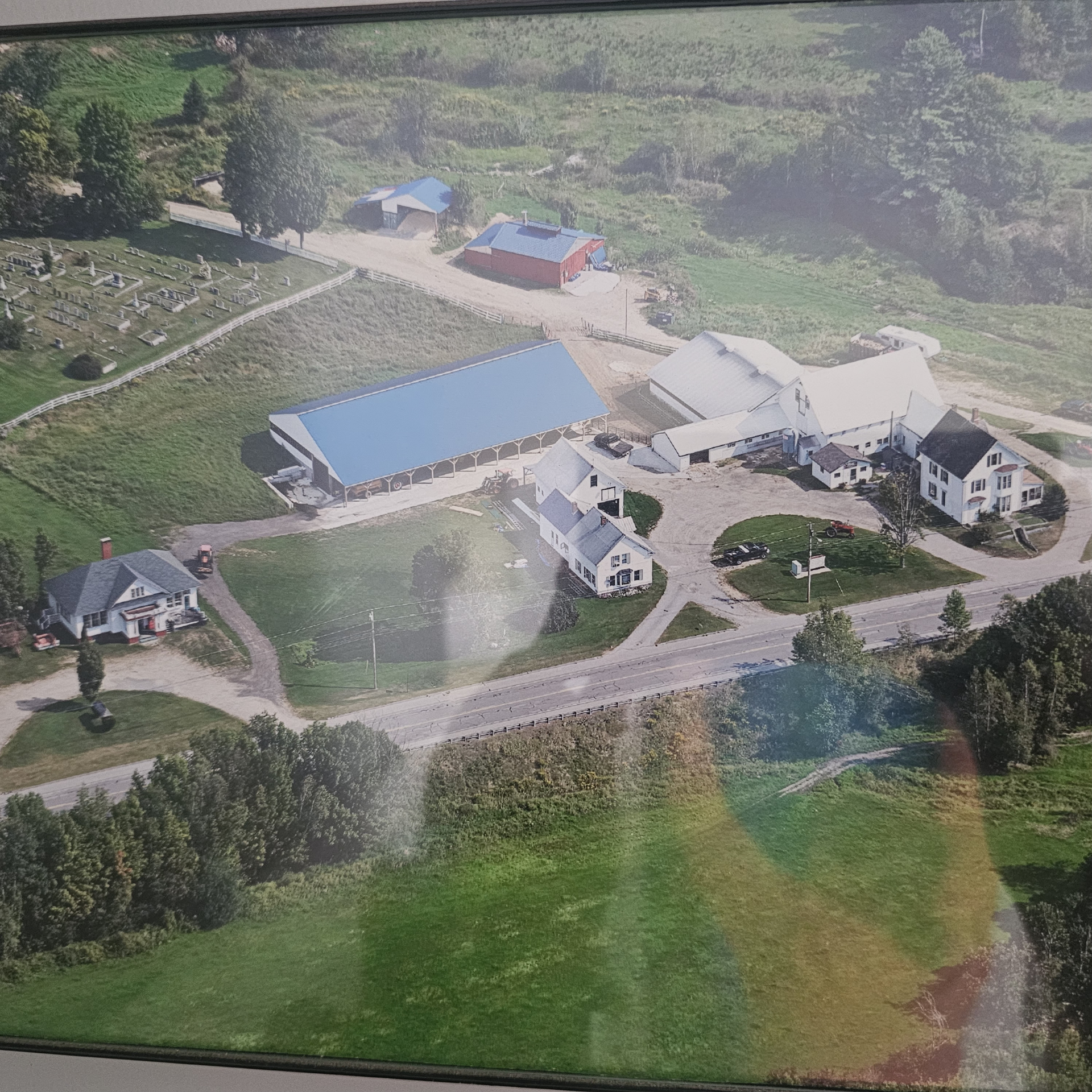 An aerial view of a farm property featuring a large blue-roofed barn, smaller outbuildings, and a farmhouse by a road.