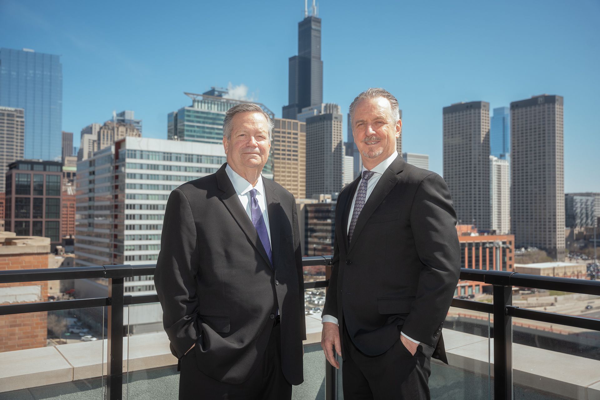 Two men in suits standing on a rooftop with a city skyline behind them.