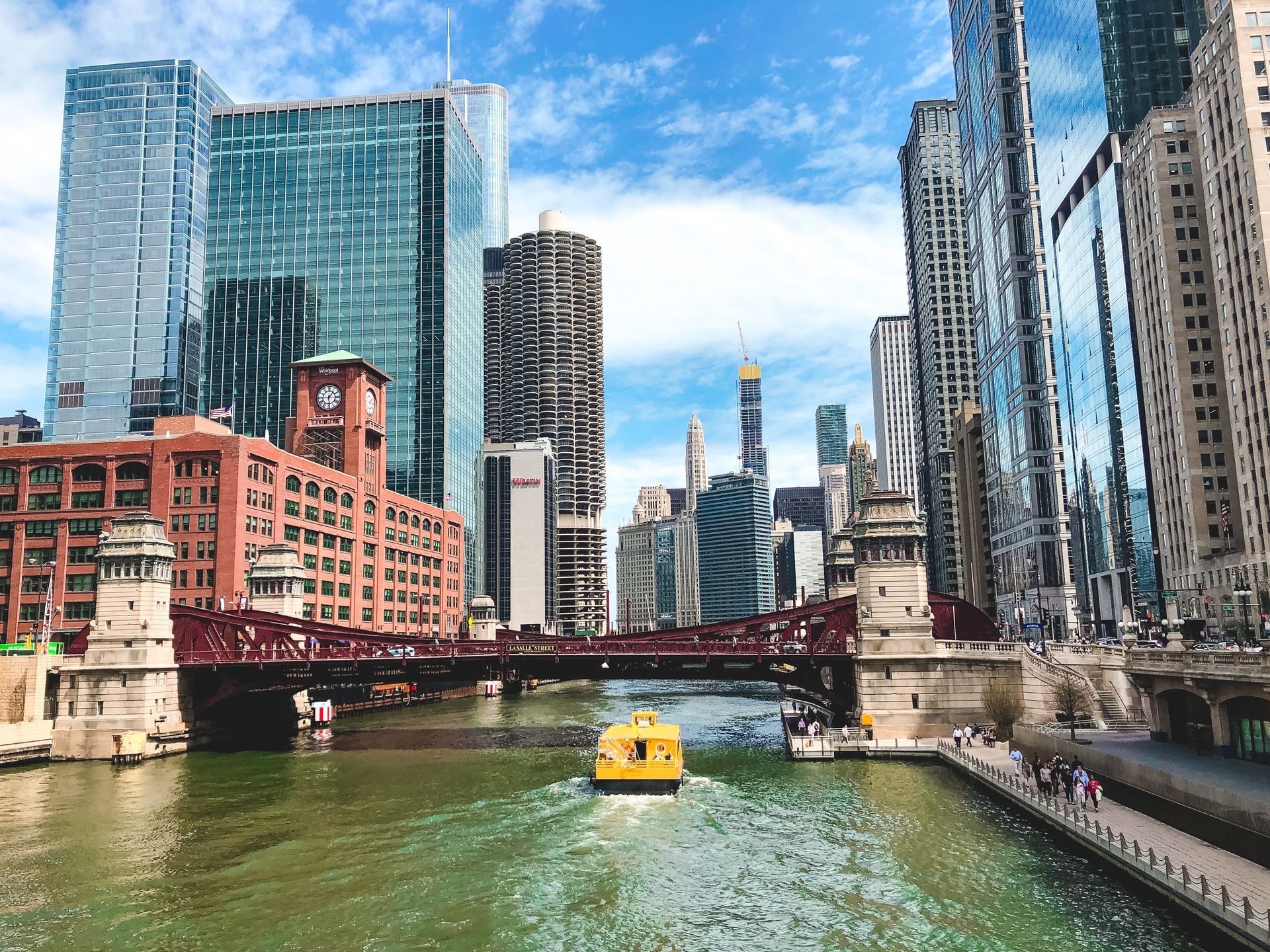 Chicago River with a yellow water taxi passing under a bridge, framed by downtown skyscrapers on a sunny day