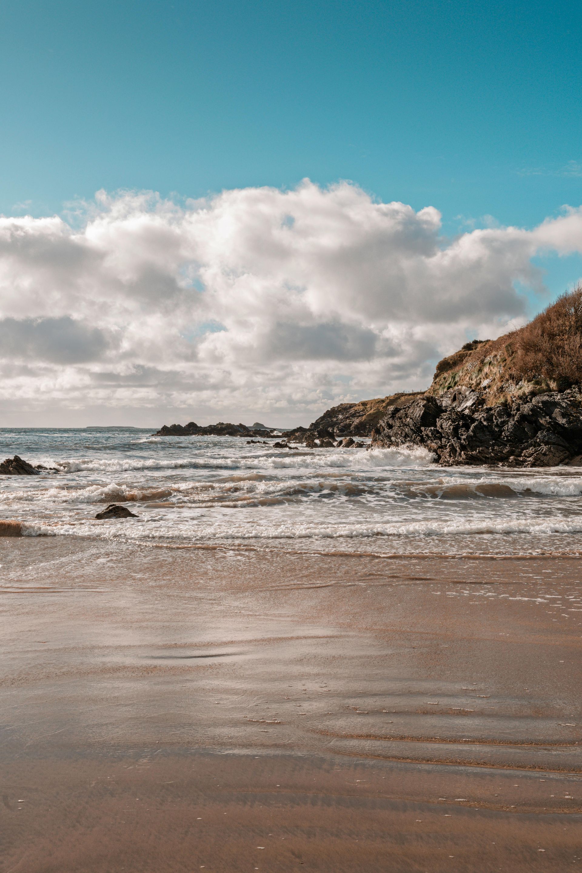 Sandy beach with gentle waves, rocky shoreline, and cloudy blue sky