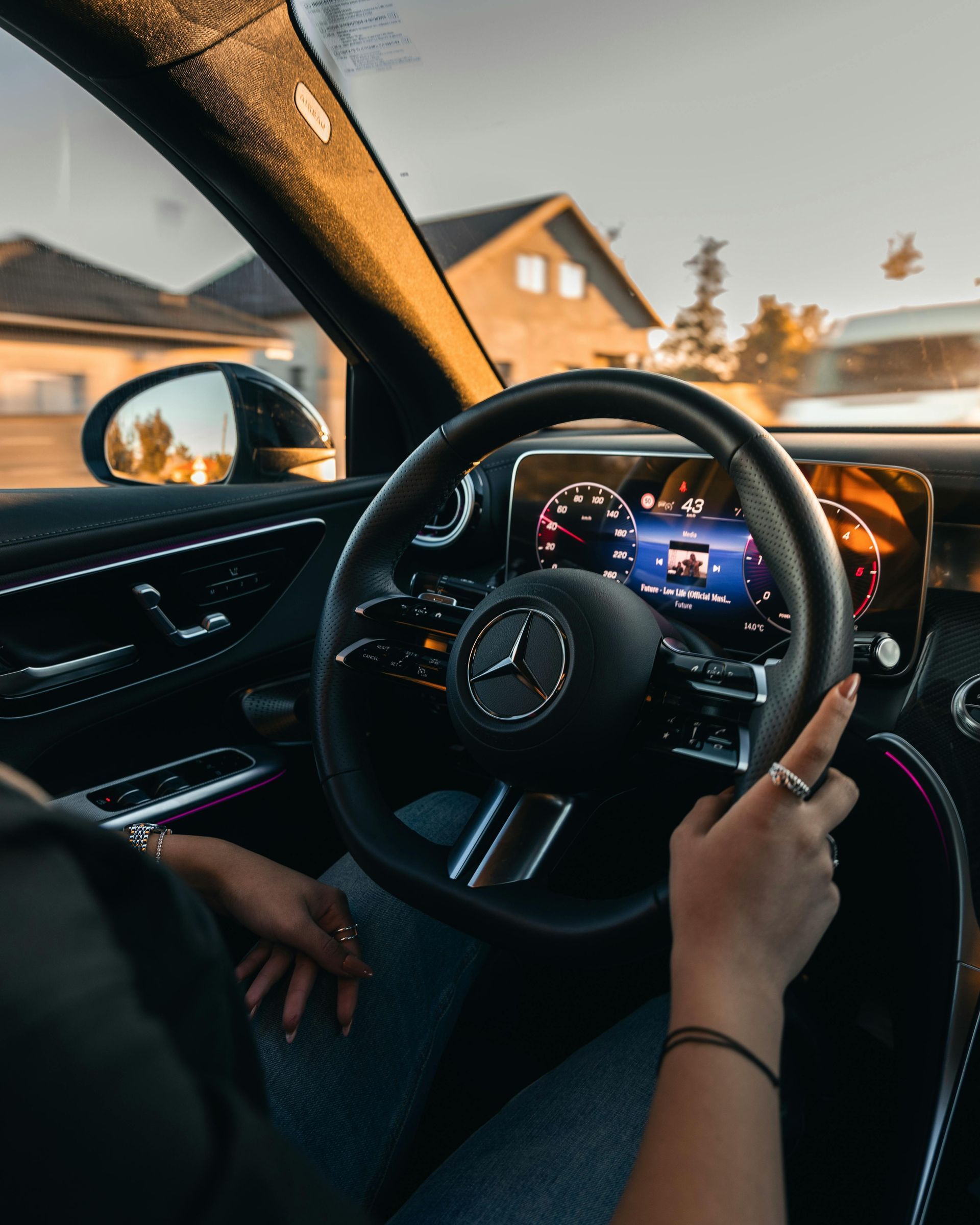 Mercedes steering wheel and dashboard viewed from inside a car at sunset, with a hand on the wheel.