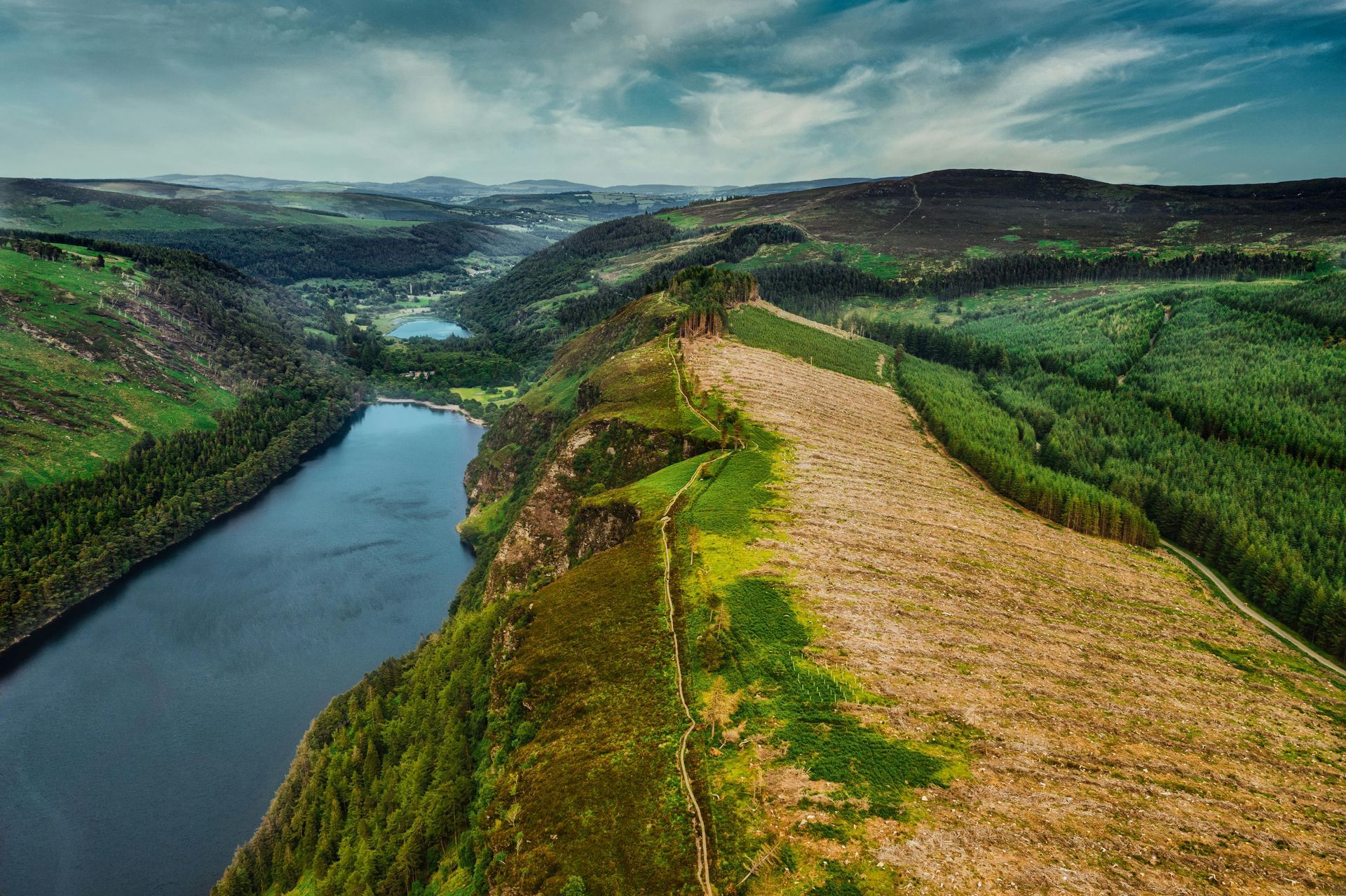 Aerial view of a river beside green hills and a tan cleared ridge under cloudy skies