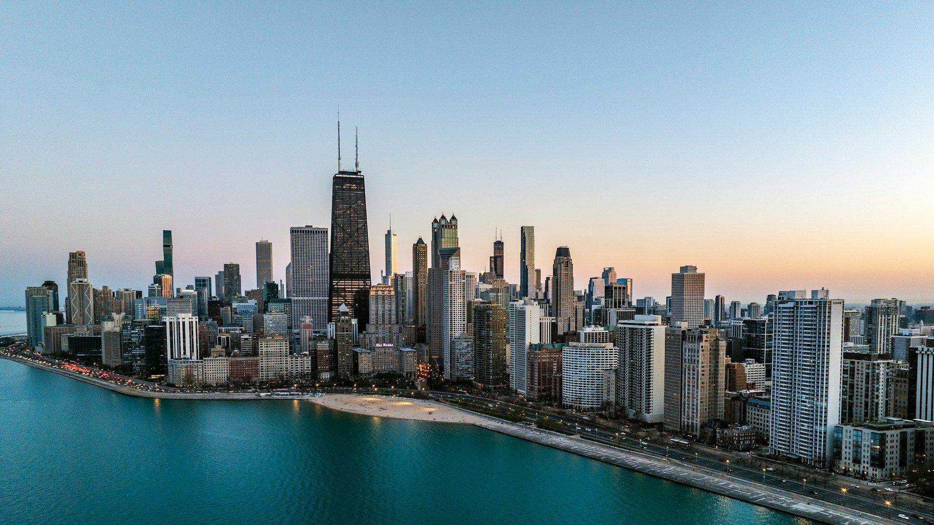 City skyline at dusk beside a waterfront, with tall modern buildings and calm blue water.