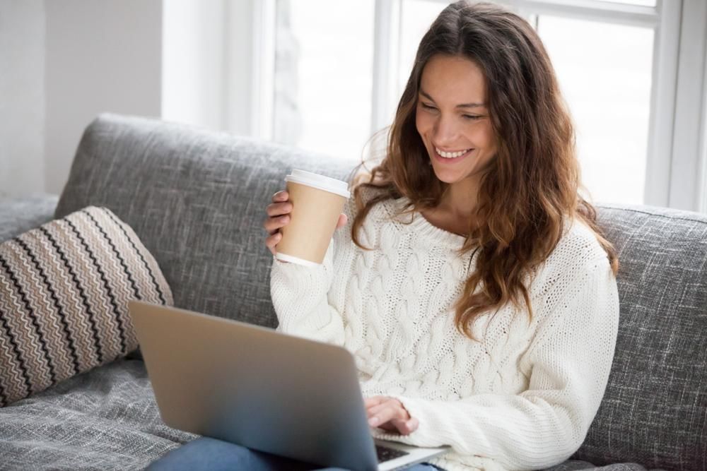 A Woman Is Sitting On A Couch Using A Laptop Computer While Holding A Cup Of Coffee — Howells & Co Legal & Conveyancing In Tamworth, NSW