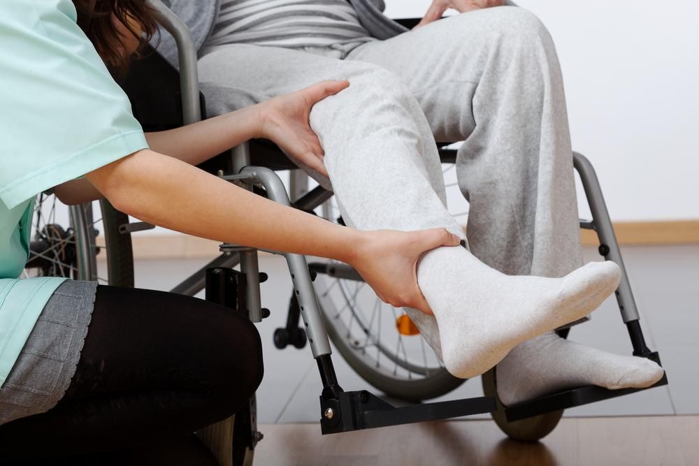 A Woman Is Helping A Man In A Wheelchair Stretch His Leg — Howells & Co Legal & Conveyancing In Cessnock, NSW